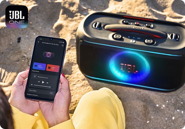 JBL ONE 앱 로고가 좌측 상단에 나와있고 모래 위에 JBL PARTYBOX 스피커가 있습니다. 그 앞에서 앱으로 스피커를 제어하는 모습이 연출되어 있습니다.