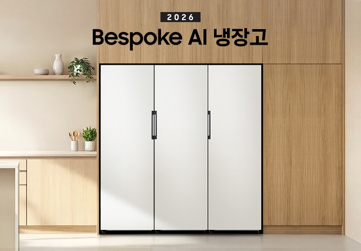 2026 Bespoke AI 냉장고. 우트톤 냉장고장에 냉장고가 설치되어 있습니다.  냉장고 좌측에는 베이지톤 하부장과 수전이 있으며 아일랜드 식탁이 연결되어 있습니다. 냉장고 바로 좌측 하부장 위에 수저를 담은 통과 화분이 있으며, 아일랜드 식탁 앞쪽에는 등받이 없는 우드톤 의자가 두 개 놓여 있습니다. 식탁 위에는 샐러드가 담긴 보울과 오렌지 주스가 담긴 컵이 놓여있습니다. 냉장고장의 바로 좌측 상단에는 우드톤 선반이 있으며 선반 위에 오브제 스타일의 그릇, 물병과 화분이 놓여있는 연출된 이미지입니다. 