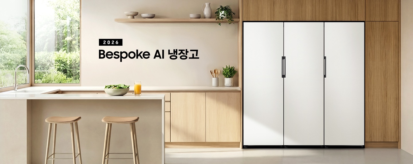 2026 Bespoke AI 냉장고. 우트톤 냉장고장에 냉장고가 설치되어 있습니다.  냉장고 좌측에는 베이지톤 하부장과 수전이 있으며 아일랜드 식탁이 연결되어 있습니다. 냉장고 바로 좌측 하부장 위에 수저를 담은 통과 화분이 있으며, 아일랜드 식탁 앞쪽에는 등받이 없는 우드톤 의자가 두 개 놓여 있습니다. 식탁 위에는 샐러드가 담긴 보울과 오렌지 주스가 담긴 컵이 놓여있습니다. 냉장고장의 바로 좌측 상단에는 우드톤 선반이 있으며 선반 위에 오브제 스타일의 그릇, 물병과 화분이 놓여있는 연출된 이미지입니다. 