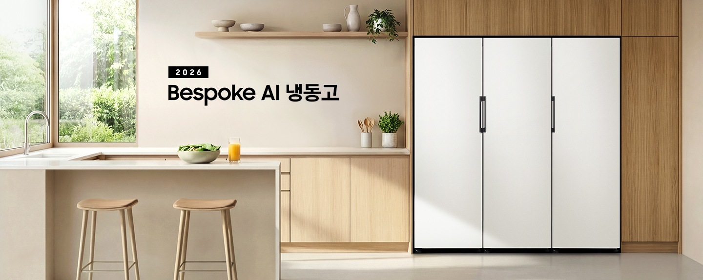 2026 Bespoke AI 냉동고. 우트톤 냉장고장에 냉동고가 설치되어 있습니다.  냉동고 좌측에는 베이지톤 하부장과 수전이 있으며 아일랜드 식탁이 연결되어 있습니다. 냉동고 바로 좌측 하부장 위에 수저를 담은 통과 화분이 있으며, 아일랜드 식탁 앞쪽에는 등받이 없는 우드톤 의자가 두 개 놓여 있습니다. 식탁 위에는 샐러드가 담긴 보울과 오렌지 주스가 담긴 컵이 놓여있습니다. 냉장고장의 바로 좌측 상단에는 우드톤 선반이 있으며 선반 위에 오브제 스타일의 그릇, 물병과 화분이 놓여있는 연출된 이미지입니다. 