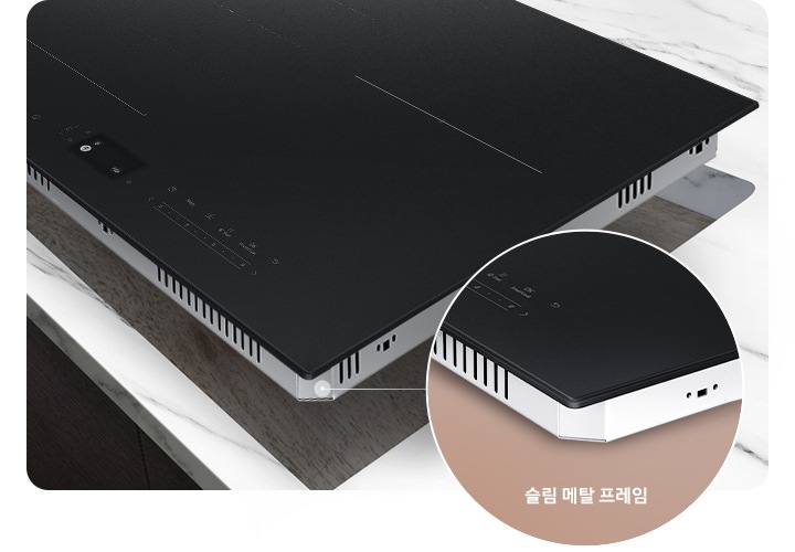 진한 갈색의 마루가 깔린 바닥이 보이고 어두운 갈색 패널의 주방 위 흰색 대리석 상판이 깔린 주방 테이블이 있습니다. 그 위에 인덕션을 설치할 부위가 타공되어 있고, 타공 된 위치 조금 위로 인덕션이 놓여 있습니다. 인덕션 오른쪽 아래에는 닷 모양의 표시와 함께 원형으로 프레임 부분이 확대되어 있습니다. 확대된 프레임 하단에는 슬림 메탈 프레임이라는 문구가 적혀 있습니다.