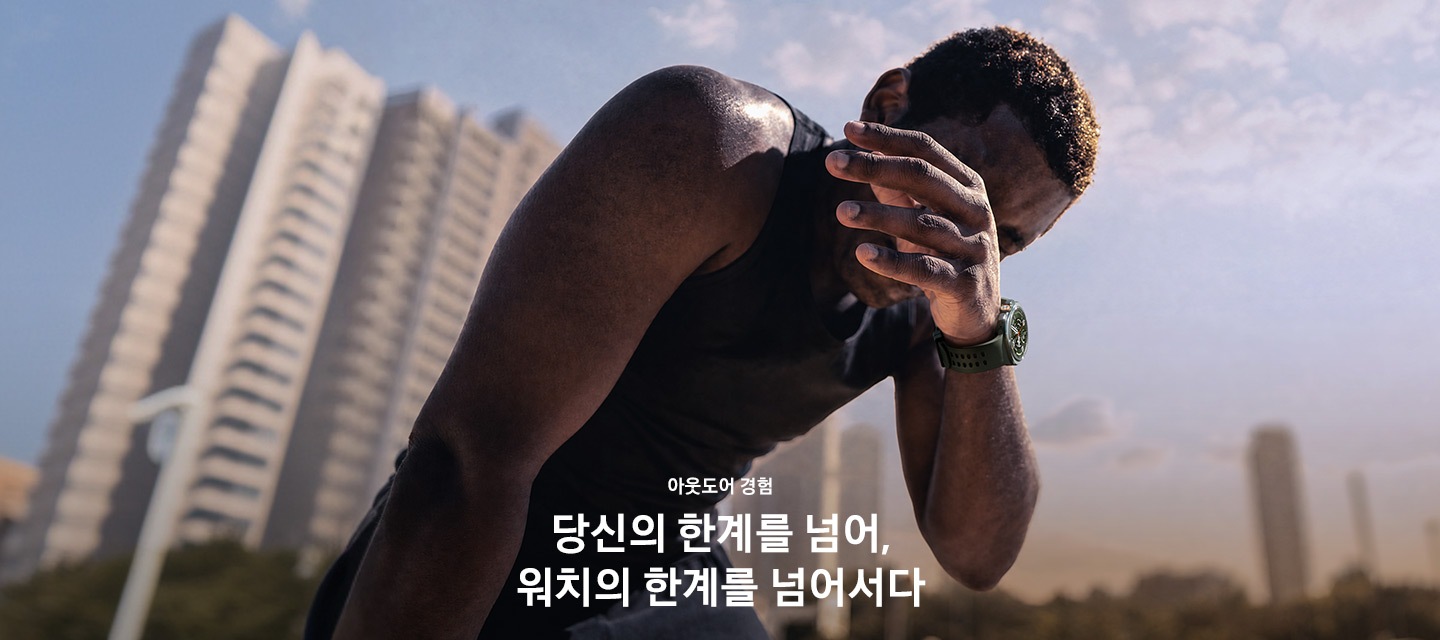 스웨트셔츠를 입고 밝은 대낮에 땀을 흘리는 남성의 모습입니다. 손목에는 올리브 색상의 마린 밴드를 장착한 티타늄 실버 색상의 갤럭시 워치 울트라2를 착용하고 있으며 운동 후에 손으로 얼굴의 땀을 닦고 있습니다.