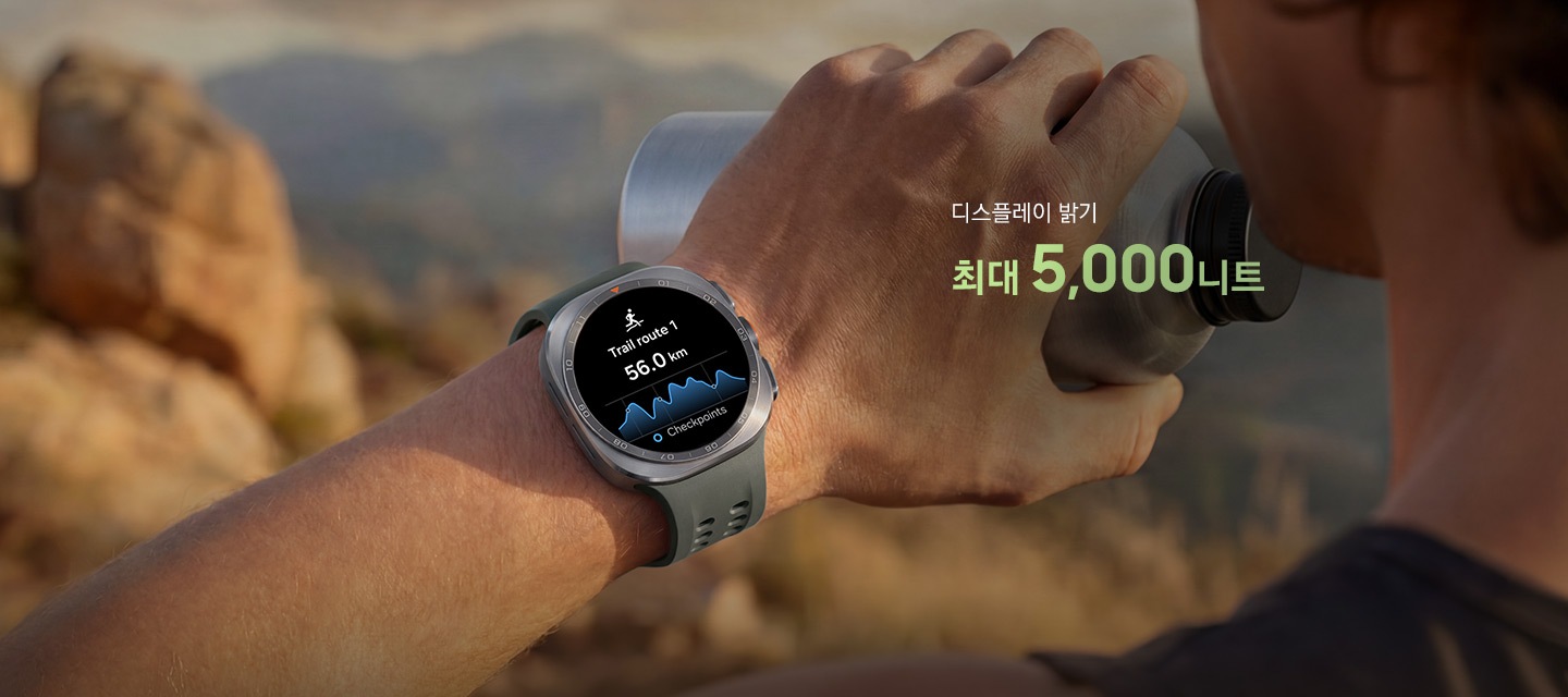 한 남성이 바위가 많은 산길을 걷고 있고, 물통을 든 손에는 올리브 색상의 마린 밴드를 장착한 티타늄 실버 색상의 갤럭시 워치 울트라2를 착용한 모습입니다. 화면에는 거리가 표시된 트레일 경로가 나타나 있어, 트레일 러닝이나 등반 시 까다로운 야외 환경에서도 신뢰할 수 있는 길 안내를 제공하는 점을 표현합니다.