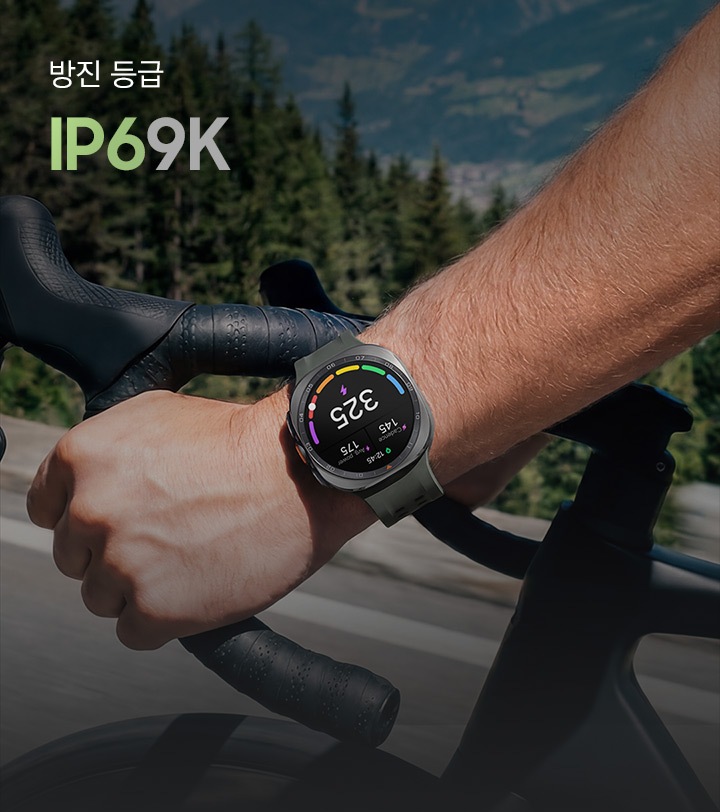 자전거 라이더가 산악 도로에서 자전거를 타고 있는 모습입니다. 라이더는 올리브 색상의 마린 밴드를 장착한 티타늄 실버 색상의 갤럭시 워치 울트라2를 착용하고 있으며, 워치 화면에는 케이던스, 개인 맞춤형 FTP(젖산 역치 파워), 평균 파워가 표시되어 있습니다.&nbsp;