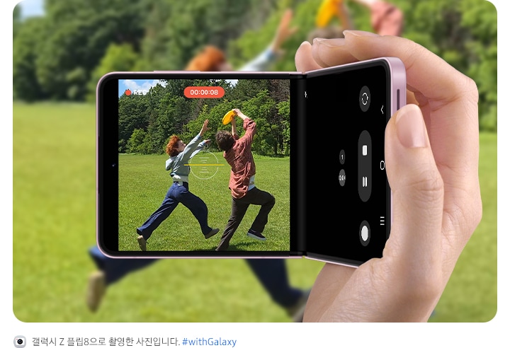 갤럭시 Z 플립8을 캠코더처럼 손에 들고 야외에서 즐겁게 놀고 있는 두 친구를 촬영하는 모습입니다. 수평 고정 슈퍼 스테디의 수평 잠금 기능 덕분에 영상이 흔들림 없이 안정적으로 유지되는 것을 강조하고 있습니다. 