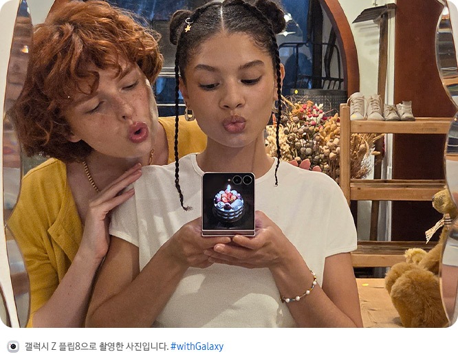 두 사람이 거울 셀피를 촬영하는 모습입니다. 한 사람이 갤럭시 Z 플립8을 플렉스모드 상태로 들고 있고, 사용자 지정 사진으로 맞춤 설정된 커버 디스플레이가 나타납니다.