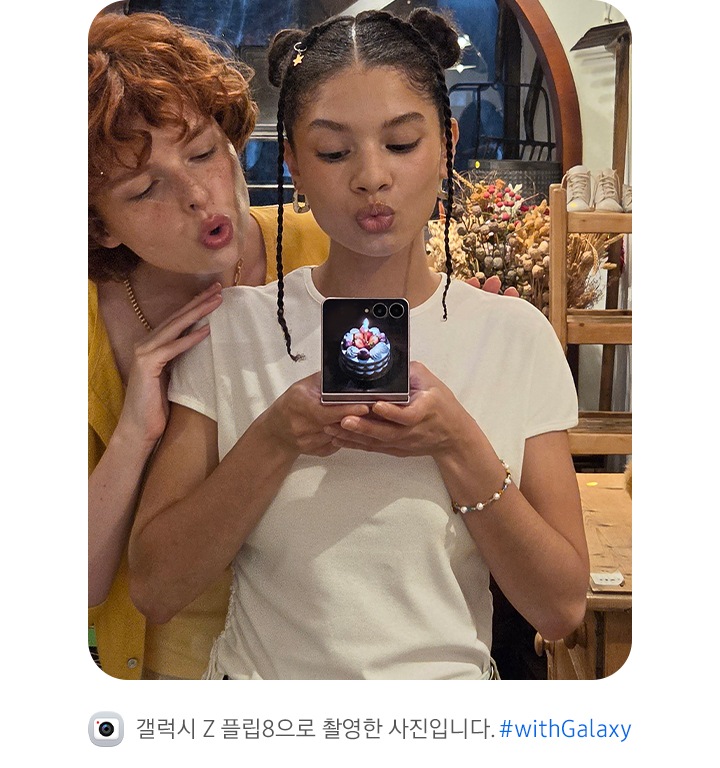 두 사람이 거울 셀피를 촬영하는 모습입니다. 한 사람이 갤럭시 Z 플립8을 플렉스모드 상태로 들고 있고, 사용자 지정 사진으로 맞춤 설정된 커버 디스플레이가 나타납니다.