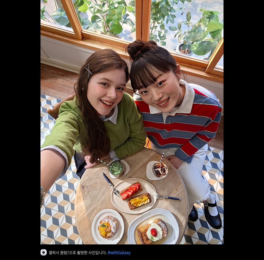 갤럭시 퀀텀7의 전면 카메라로 위에서 아래를 향해 촬영한 사진입니다. 밝은 조명의 카페에서 브런치를 즐기고 있는 두 여성의 셀피 사진입니다. 슈퍼 HDR 덕분에 사진의 디테일이 생생하고 색감이 풍부합니다. 하단에는 카메라 아이콘과 함께 '갤럭시 퀀텀7으로 촬영한 영상입니다' 문구와 #with_galaxy 해시태그가 표시되어 있습니다.
