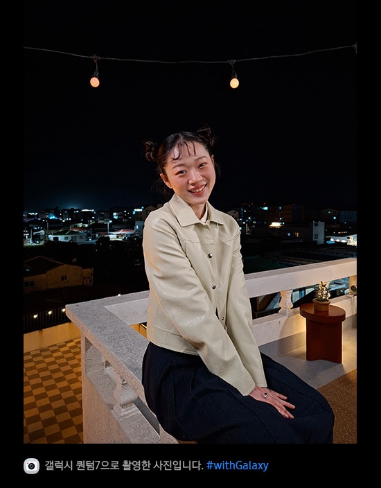 갤럭시 퀀텀7으로 촬영한 밤에 야외 테라스에 기대어 있는 젊은 여성의 사진입니다. 최적화된 나이토그래피 덕분에 사진의 디테일이 밝고 선명합니다. 하단에는 카메라 아이콘과 함께 '갤럭시 퀀텀7으로 촬영한 영상입니다' 문구와 #with_galaxy 해시태그가 표시되어 있습니다.