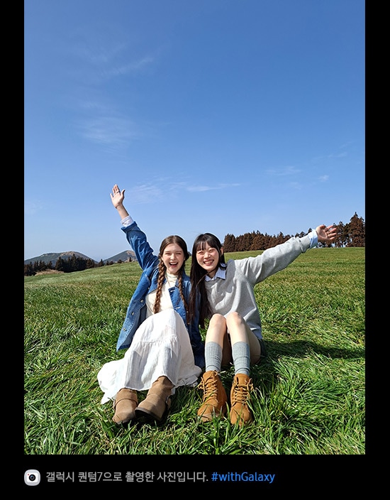 갤럭시 퀀텀7의 광각 카메라로 촬영한 사진입니다. 푸른 하늘을 배경으로 탁 트인 잔디밭 한가운데에 두 여성이 앉아 있습니다. 사진의 디테일이 밝고 선명하며 화질이 향상되었습니다. 하단에는 카메라 아이콘과 함께 '갤럭시 퀀텀7으로 촬영한 영상입니다' 문구와 #with_galaxy 해시태그가 표시되어 있습니다.