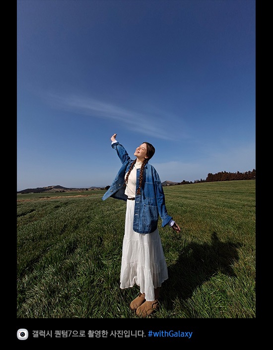 갤럭시 퀀텀7의 초광각 카메라로 촬영한 사진입니다. 푸른 하늘을 배경으로 탁 트인 잔디밭 한가운데 여성이 서 있습니다. 사진의 디테일이 선명하고 생생하며 화질이 향상되었습니다. 하단에는 카메라 아이콘과 함께 '갤럭시 퀀텀7으로 촬영한 영상입니다' 문구와 #with_galaxy 해시태그가 표시되어 있습니다.