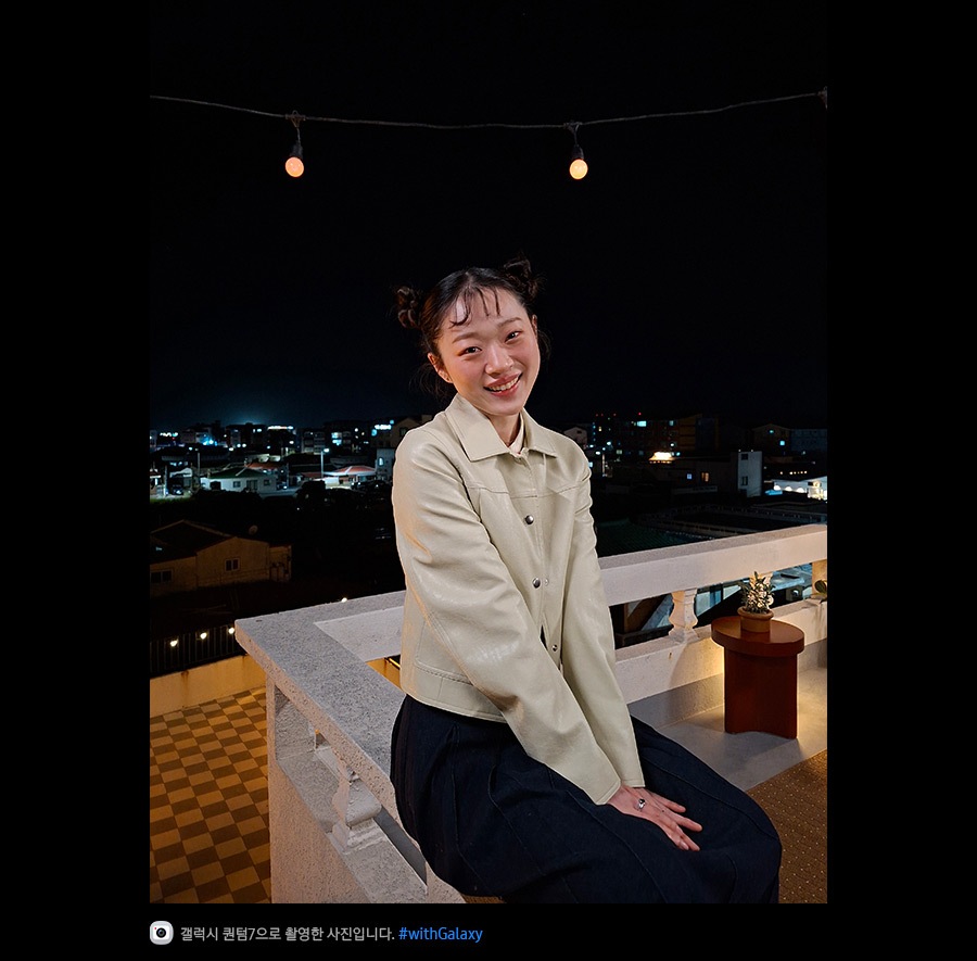 갤럭시 퀀텀7으로 촬영한 밤에 야외 테라스에 기대어 있는 젊은 여성의 사진입니다. 최적화된 나이토그래피 덕분에 사진의 디테일이 밝고 선명합니다. 하단에는 카메라 아이콘과 함께 '갤럭시 퀀텀7으로 촬영한 영상입니다' 문구와 #with_galaxy 해시태그가 표시되어 있습니다.