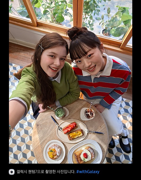 갤럭시 퀀텀7의 전면 카메라로 위에서 아래를 향해 촬영한 사진입니다. 밝은 조명의 카페에서 브런치를 즐기고 있는 두 여성의 셀피 사진입니다. 슈퍼 HDR 덕분에 사진의 디테일이 생생하고 색감이 풍부합니다. 하단에는 카메라 아이콘과 함께 '갤럭시 퀀텀7으로 촬영한 영상입니다' 문구와 #with_galaxy 해시태그가 표시되어 있습니다.
