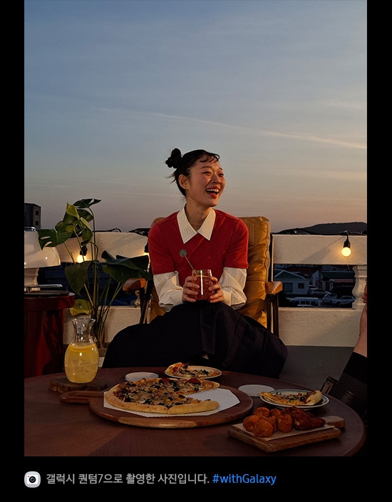 갤럭시 퀀텀7으로 촬영한 어두운 환경의 야외 테라스에서 미소 짓고 있는 여성의 사진입니다. 사진의 디테일이 밝고 선명합니다. 하단에는 카메라 아이콘과 함께 '갤럭시 퀀텀7으로 촬영한 영상입니다' 문구와 #with_galaxy 해시태그가 표시되어 있습니다.