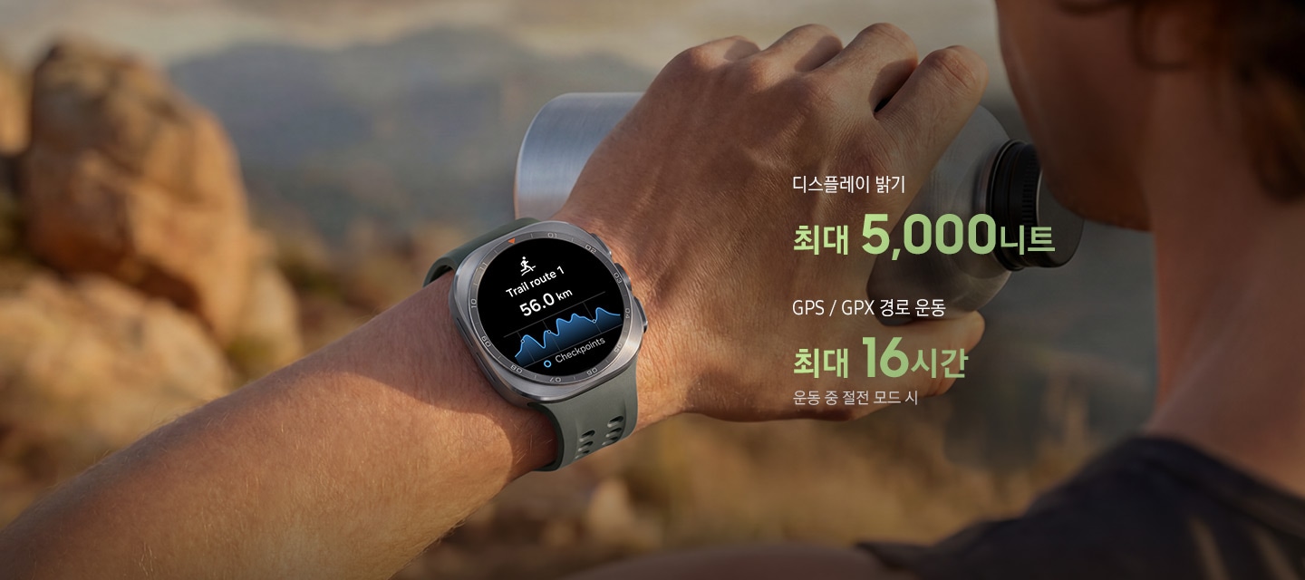 한 남성이 바위가 많은 산길을 걷고 있고, 물통을 든 손에는 올리브 색상의 마린 밴드를 장착한 티타늄 실버 색상의 갤럭시 워치 울트라2를 착용한 모습입니다. 화면에는 거리가 표시된 트레일 경로가 나타나 있어, 트레일 러닝이나 등반 시 까다로운 야외 환경에서도 신뢰할 수 있는 길 안내를 제공하는 점을 표현합니다. 이미지 내에는 '디스플레이 밝기 최대 5,000니트, GPS / GPX 경로 운동 최대 16시간 운동 중 절전 모드 시' 문구가 있습니다.