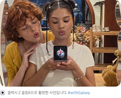 두 사람이 거울 셀피를 촬영하는 모습입니다. 한 사람이 갤럭시 Z 플립8을 플렉스모드 상태로 들고 있고, 사용자 지정 사진으로 맞춤 설정된 커버 디스플레이가 나타납니다.