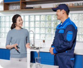[남색 옷을 입은 수리기사가 파란색 빌트인 장으로 꾸며진 주방에서 빌트인 Bésp~óké 정수기를 앞에두고 여자와 대화하는 이미지, 정수기 앞에 유리컵 물잔에 물이 따라져 있는]