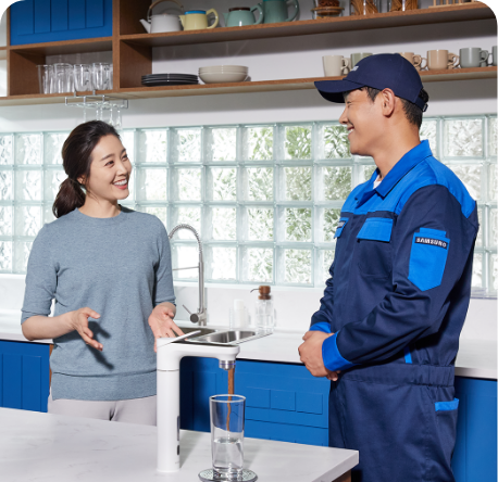 [남색 옷을 입은 수리기사가 파란색 빌트인 장으로 꾸며진 주방에서 빌트인 Bésp~óké 정수기를 앞에두고 여자와 대화하는 이미지, 정수기 앞에 유리컵 물잔에 물이 따라져 있는]
