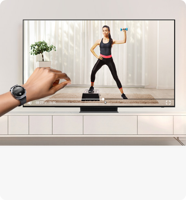 왼편에 갤럭시 워치를 착용한 손목이 보이며 중앙 뒷편의 TV 화면에는 홈트레이닝 영상이 나오고 있다