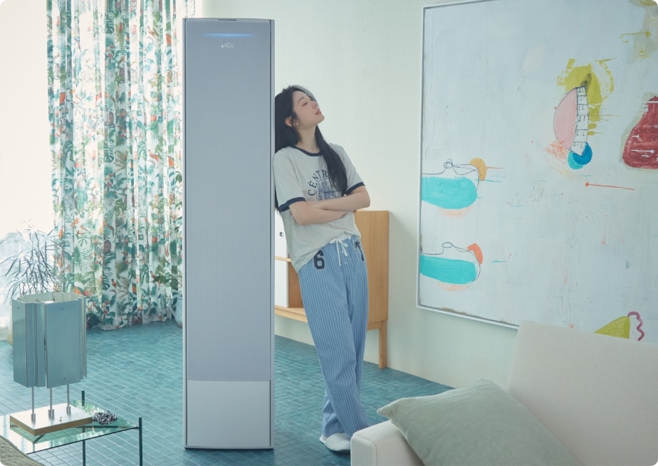 거실에서 스탠드형 에어컨 Bespoke AI 무풍콤보에 기대어 선 김연아. 편안한 캐주얼 복장과 여유로운 표정으로 실내 인테리어와 조화를 이루며, 배경에는 추상화 그림과 식물 무늬 커튼이 어우러져 있다.