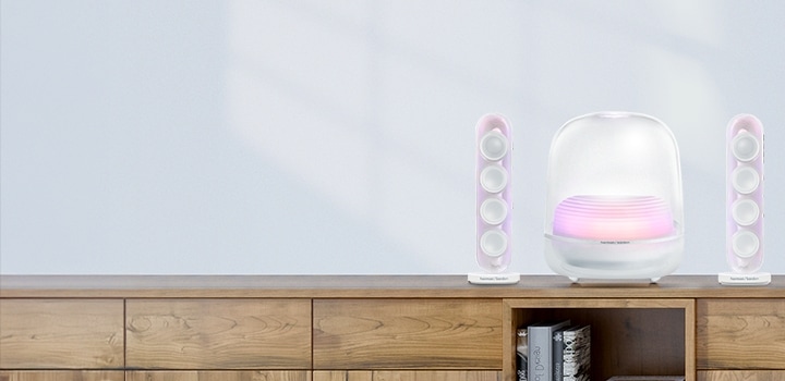 하늘색 배경 벽에 우드색 테이블 위에 놓여진 하얀색 하만카돈 SOUND STICKS 5과 화분이 놓여있는