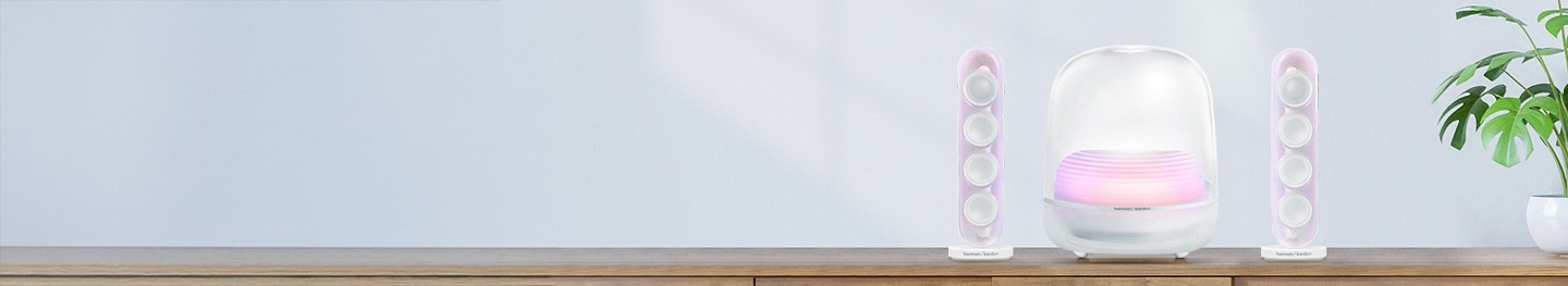 하늘색 배경 벽에 우드색 테이블 위에 놓여진 하얀색 하만카돈 SOUND STICKS 5과 화분이 놓여있는