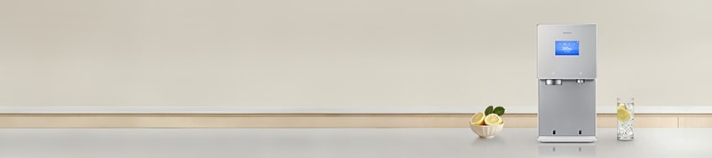 회색톤의 대리석 주방 상판 위에 2026 Bespoke AI 얼음정수기 신제품이 놓여져 있고 제품 좌측에 레몬이 담긴 그릇, 우측에는 레몬과 얼음물이 담긴 컵이 놓여있는 이미지 