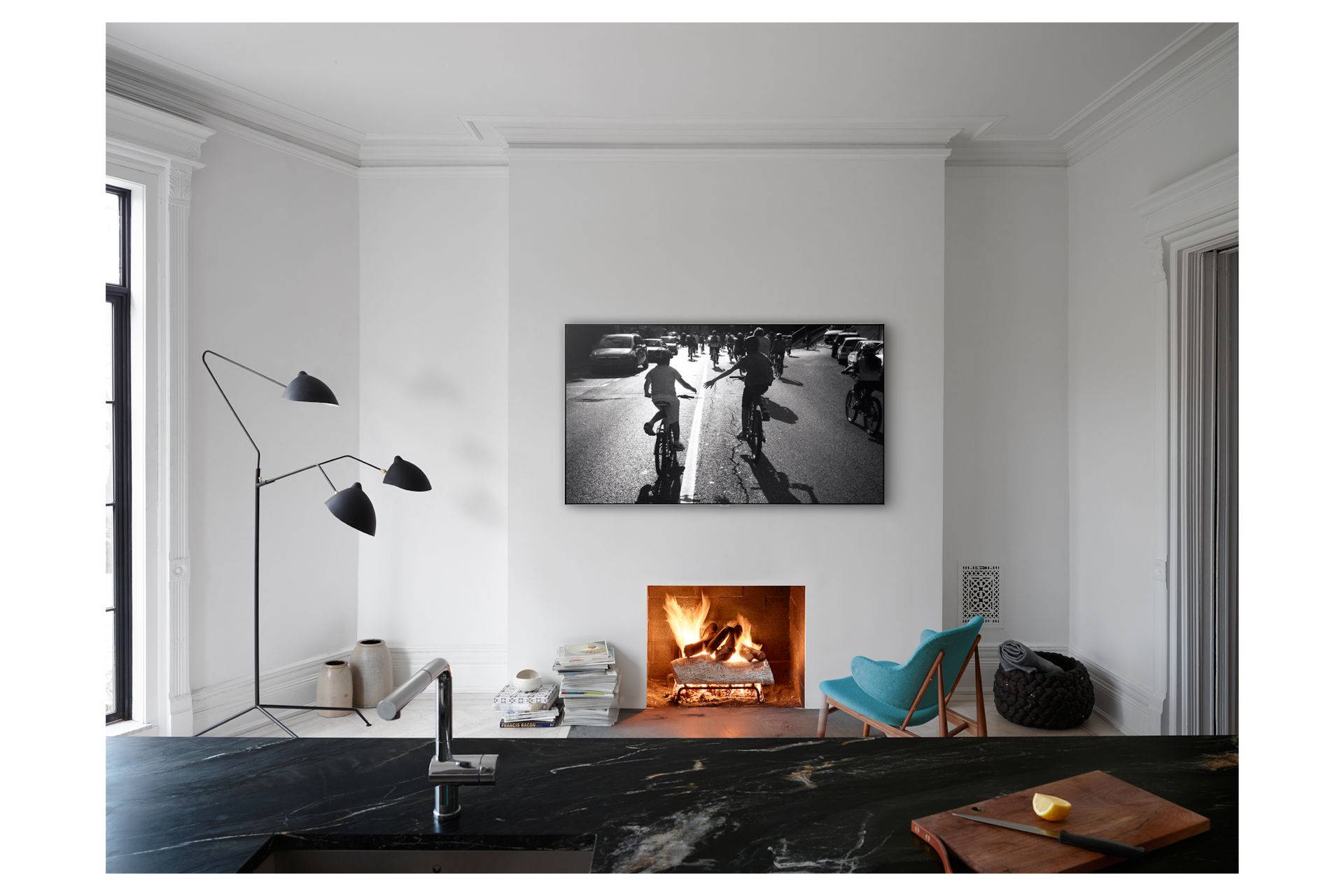 거실 전면이 보이며 벽난로가 정면에 보입니다. 벽난로 위로 QLED TV가 걸려 있습니다. 화면에는 도로위에서 자전거를 타고 있는 아이들의 모습이 흑백으로 보입니다. 