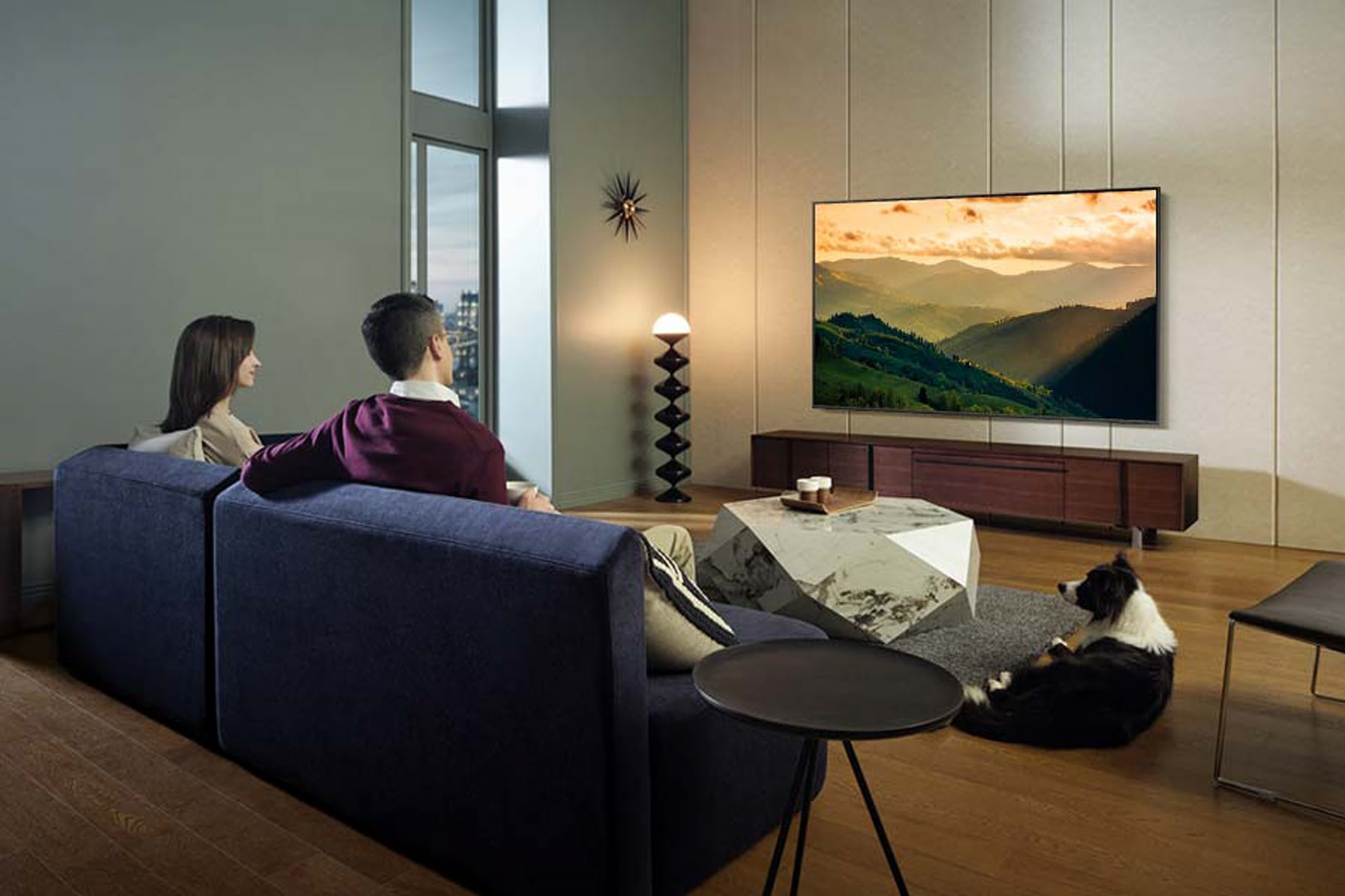 거실 벽면에 TV가 설치되어 있습니다. 화면에는 산이 보입니다. TV 앞 파란색 소파에는 남성과 여성이 앉아 있으며, 티 테이블 오른쪽에는 강아지가 엎드려 있는 라이프스타일 컷입니다.