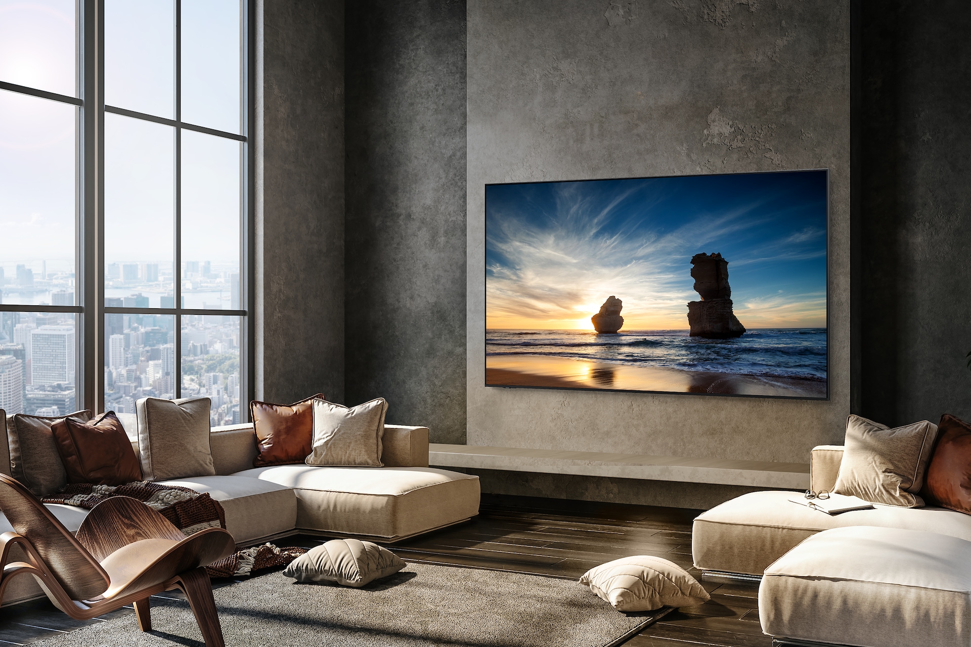 벽에 TV가 설치되어 있으며, 화면에는 노을이 지고 있는 바닷가가 보입니다. TV 왼쪽에는 창이 있습니다. TV 앞에는 소파와 쿠션들이 있는 라이프스타일 컷입니다. 
