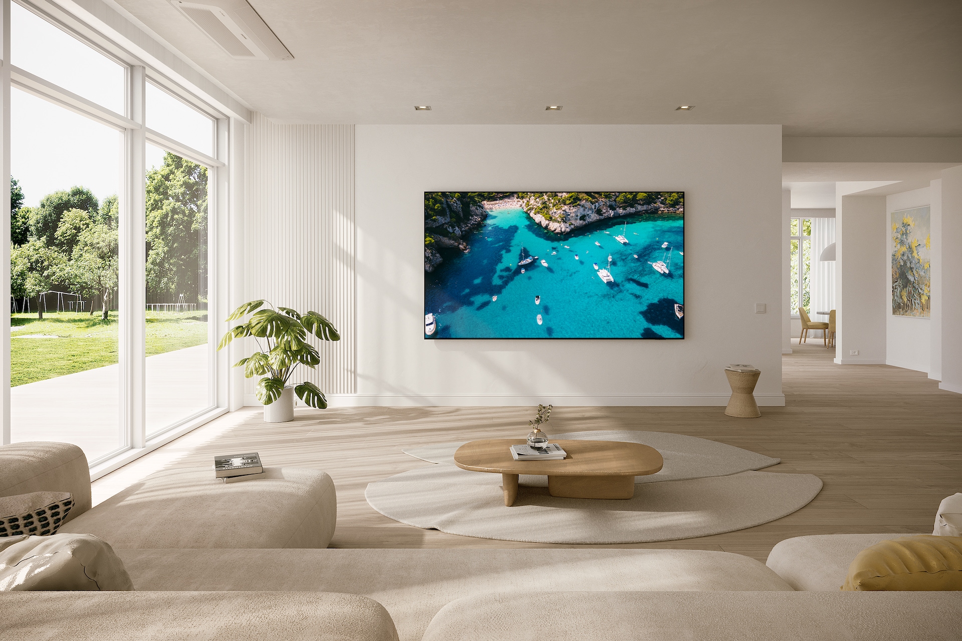 거실 벽에 TV가 설치되어 있으며, 화면에는 바다가 보입니다. TV 앞에는 테이블과 소파가 있습니다. 왼쪽 창으로는 나무가 보이는 라이프스타일컷입니다.