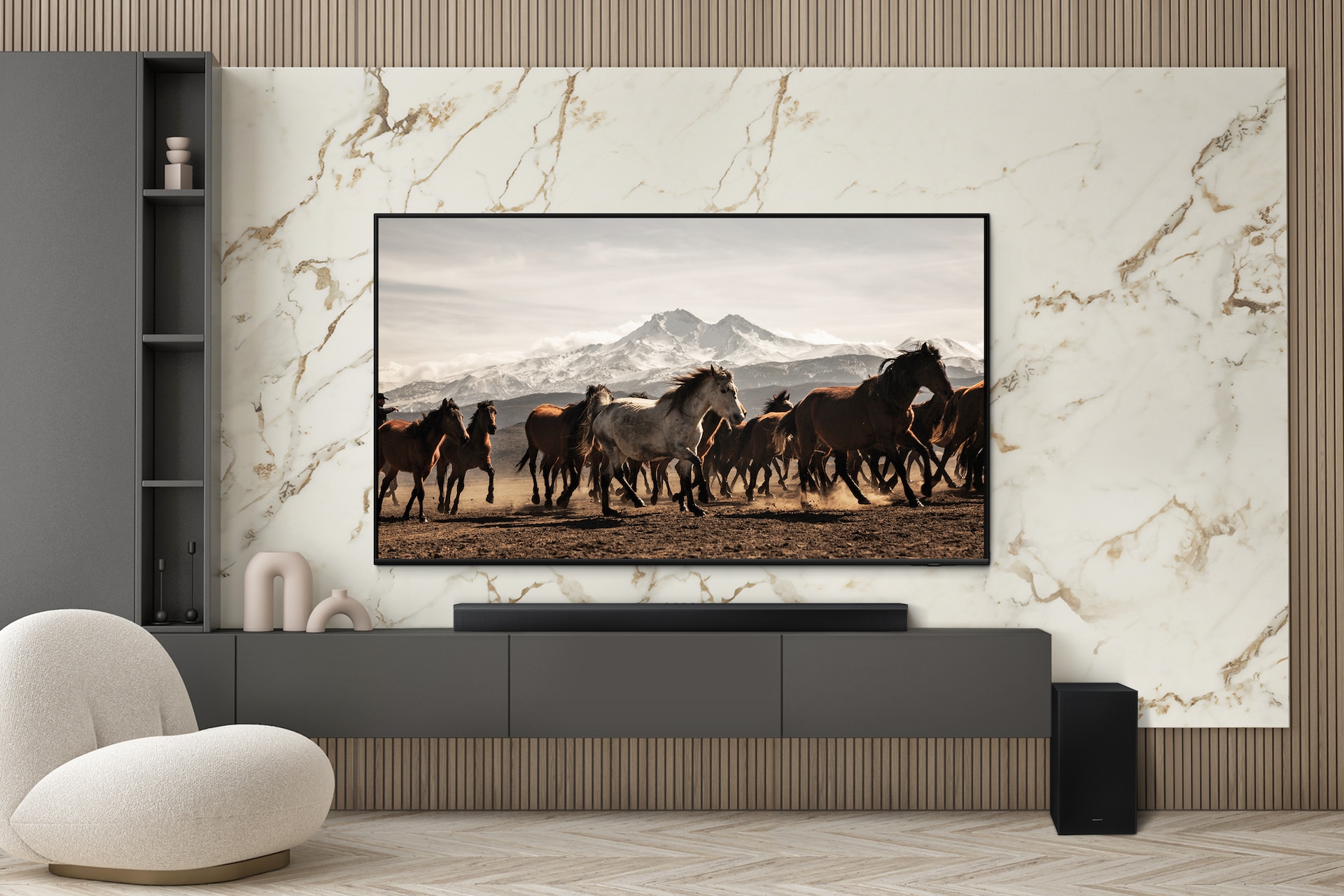 거실 벽에 TV가 설치되어 있습니다. 그 아래에는 장식장이 놓여 있으며 그 위에 사운드바가 설치되어 있습니다. 장식장 우측에는 서브우퍼가 놓여 있으며, 좌측에는 1인용 소파가 놓여 있는 라이프스타일 컷입니다.