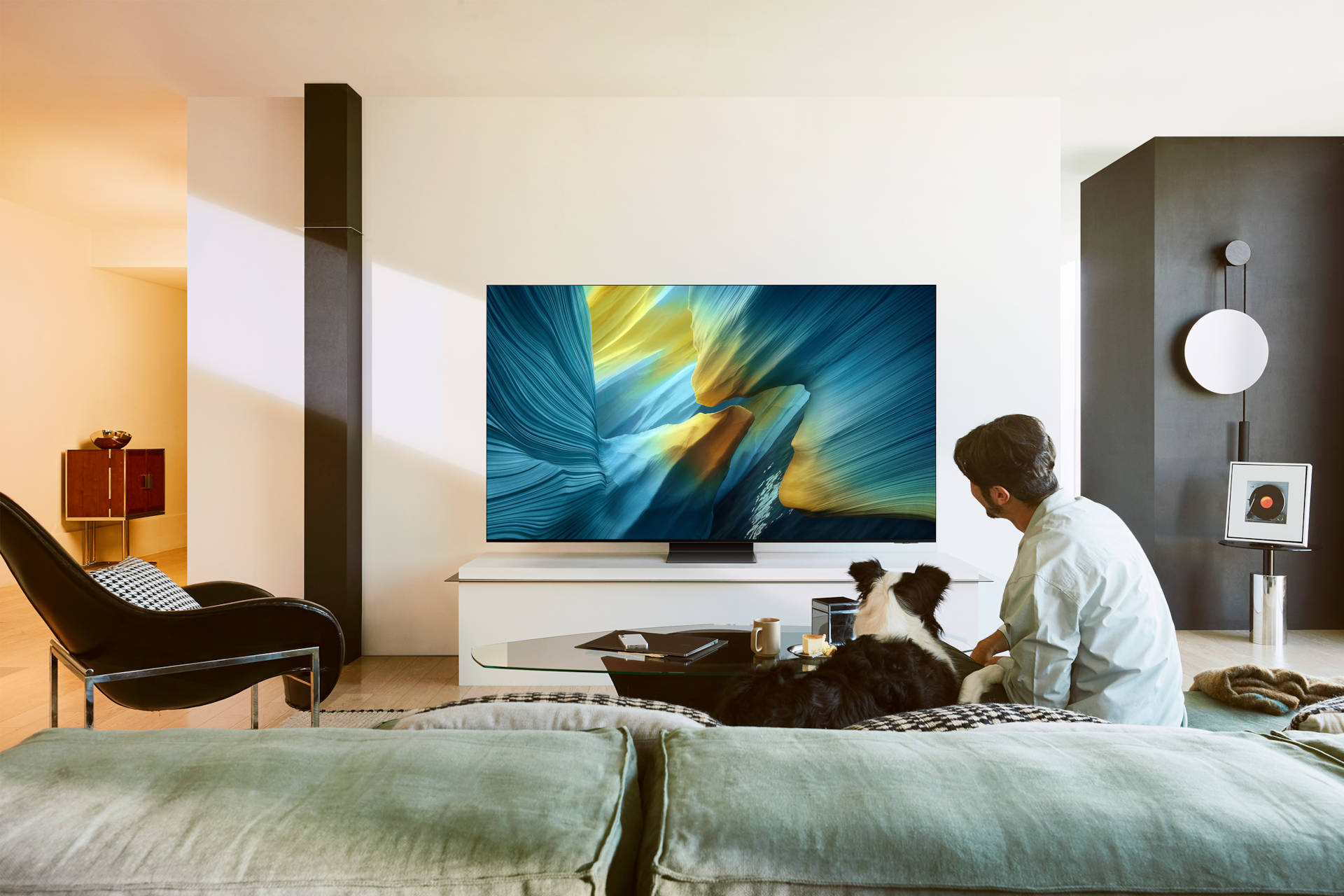 2025 OLED SF95 (138cm) 세번째 이미지이며, 거실 장식장 위에 TV가 설치되어 있습니다. 소파에는 강아지와 남성이 앉아서 TV를 보고있는 라이프스타일 컷입니다. 