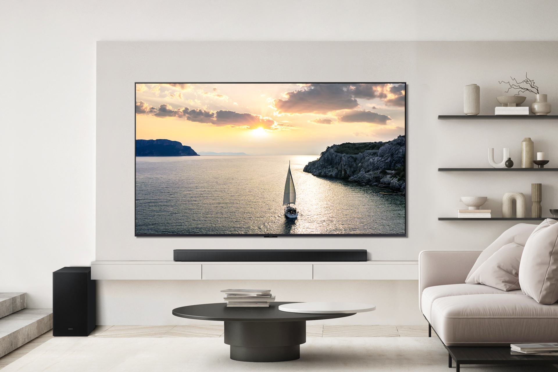 거실 벽에 TV가 설시되어 있으며, 그 하단에는 장식장이 놓여 있습니다. 장식장 위에는 사운드바가 놓여 있으며, 장식장 좌측에는 서브우퍼가 놓여 있습니다. TV 우측 벽에는 장식장 3개가 설치되어 있으며, 장식장에는 장식품들이 놓여 있습니다. TV 앞에는 테이블이 놓여 있고, 그 위에는 책들이 놓여 있습니다. 테이블 우측에는 소파가 놓여 있는 라이프스타일 컷입니다.