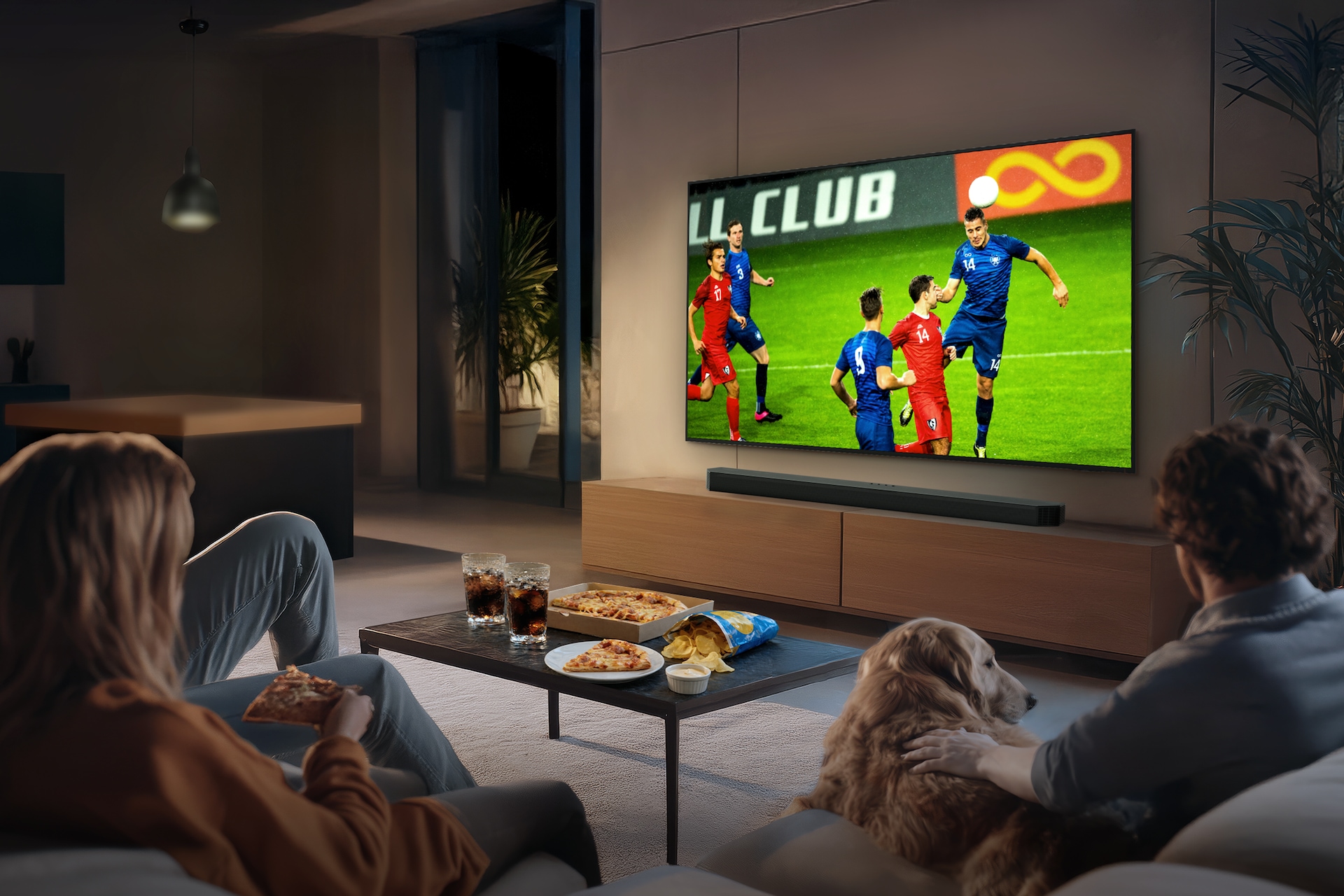 거실 벽에 TV가 설치되어 있습니다. TV 화면에는 축구 경기가 보여지고 있습니다. TV 하단에는 장식장이 있으며, 장식장 위에는 사운드바가 놓여 있습니다. TV 앞에는 테이블과 소파가 놓여 있습니다. 테이블 위에는 콜라와 피자, 과자가 놓여 있으며, 소파에는 사람들과 강아지가 앉아서 TV 화면을 보고 있습니다.