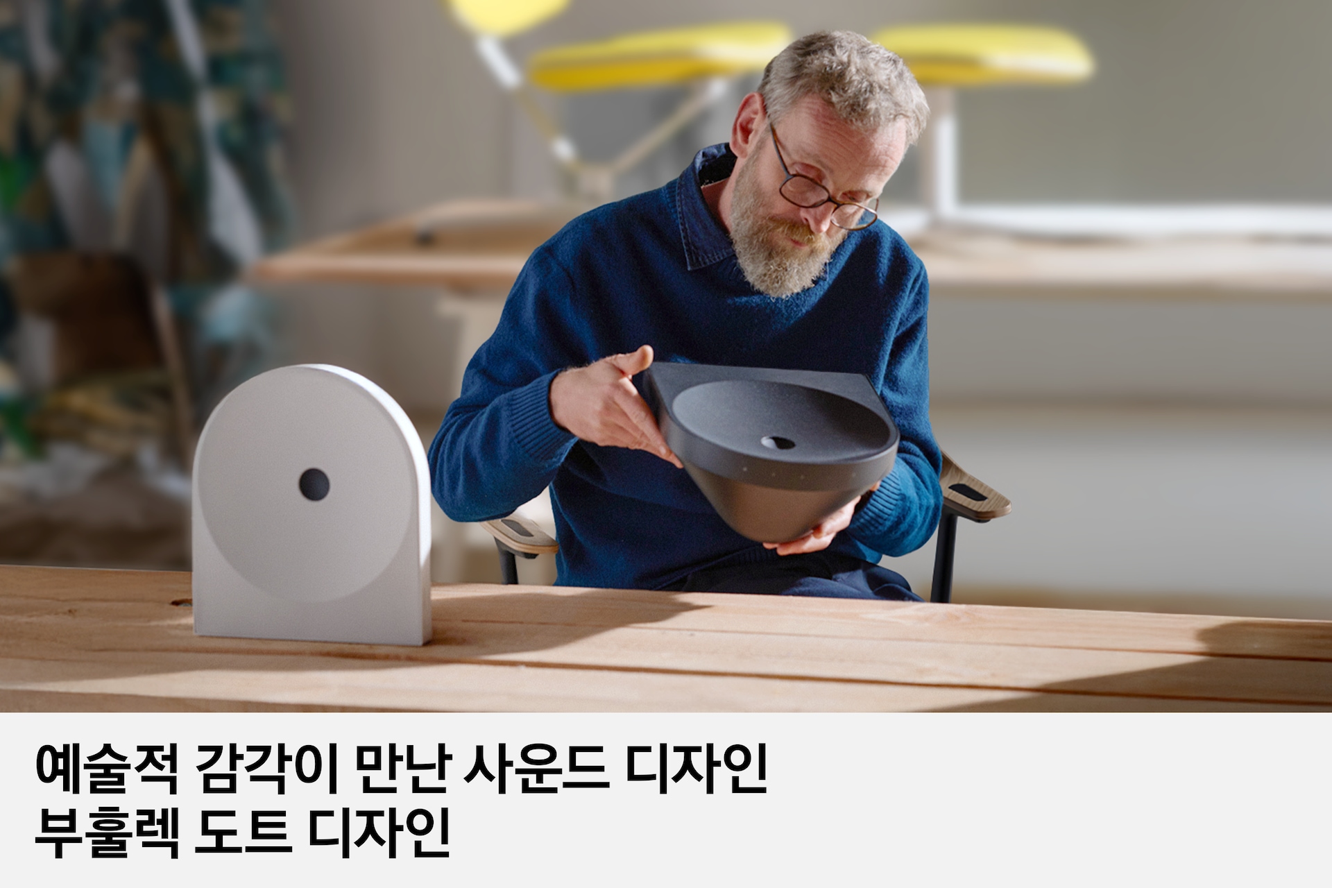 의자의 앉은 디자이너 에르완 부훌렉이 블랙 색상의 스피커를 보고 있습니다. 책상 위에는 화이트 색상의 스피커가 놓여 있습니다. 이미지 하단에는 '예술적 감각이 만난 사운드 디자인 부훌렉 도트 디자인' 문구가 기재되어 있습니다.