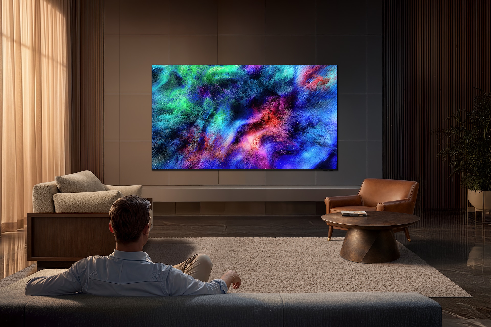 거실 벽에 TV가 설치되어 있습니다. TV 앞에는 테이블과 소파, 1인용 의자 등이 있습니다. 남성이 소파에 앉아서 TV를 보고 있습니다.