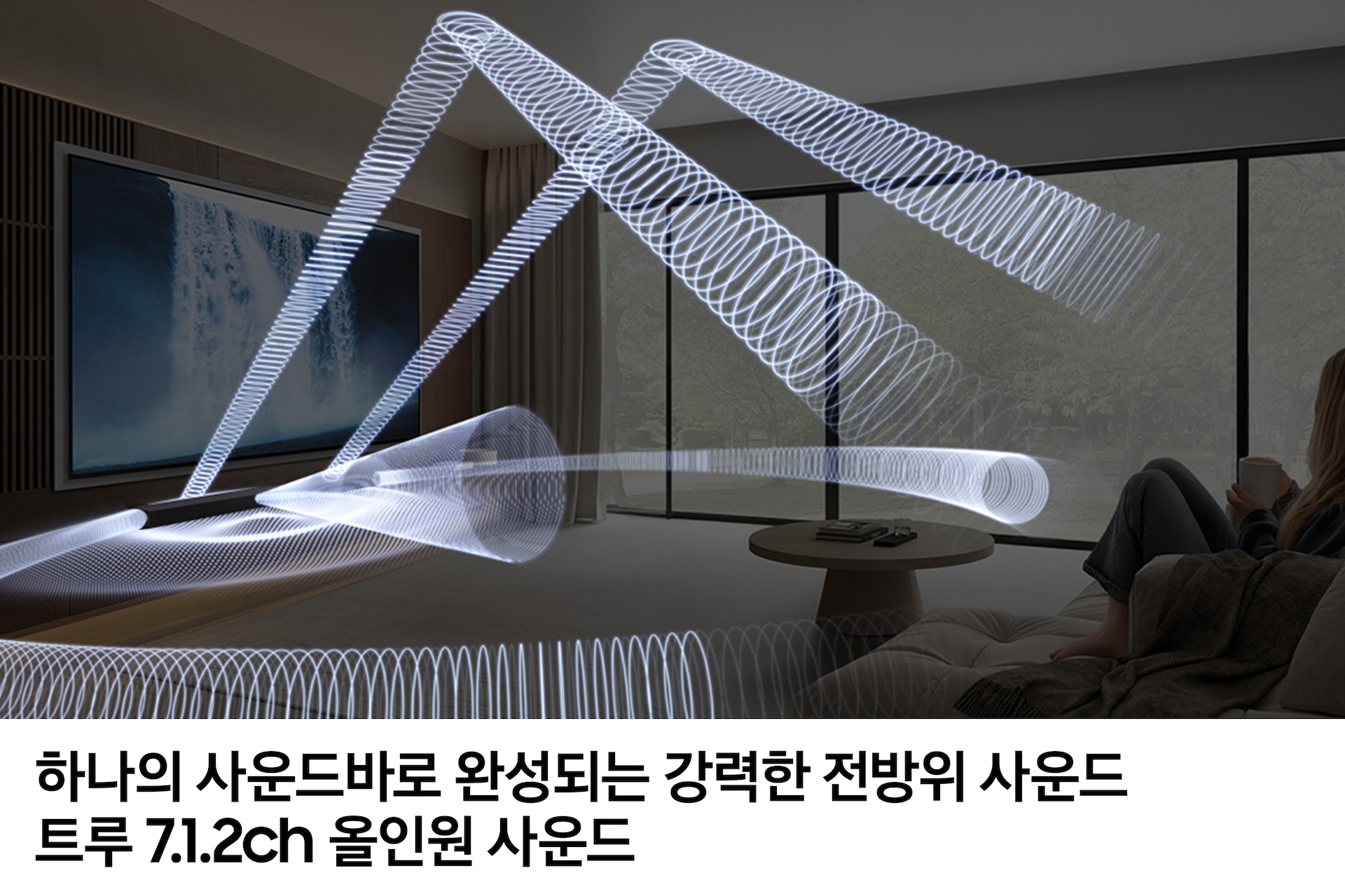 거실 벽에 TV와 사운드바 본체가 설치되어 있습니다. TV 앞에는 소파와 테이블이 놓여 있으며, 소파에는 여성이 앉아서 TV를 보고 있습니다. TV 화면에는 폭포의 모습이 보이며, TV 하단에 있는 사운드바에서 음향효과가 나오고 있습니다. 이미지 하단에는 '하나의 사운드바로 완성되는 강력한 전방위 사운드 트루 7.1.2ch 올인원 사운드' 문구가 기재되어 있습니다.
