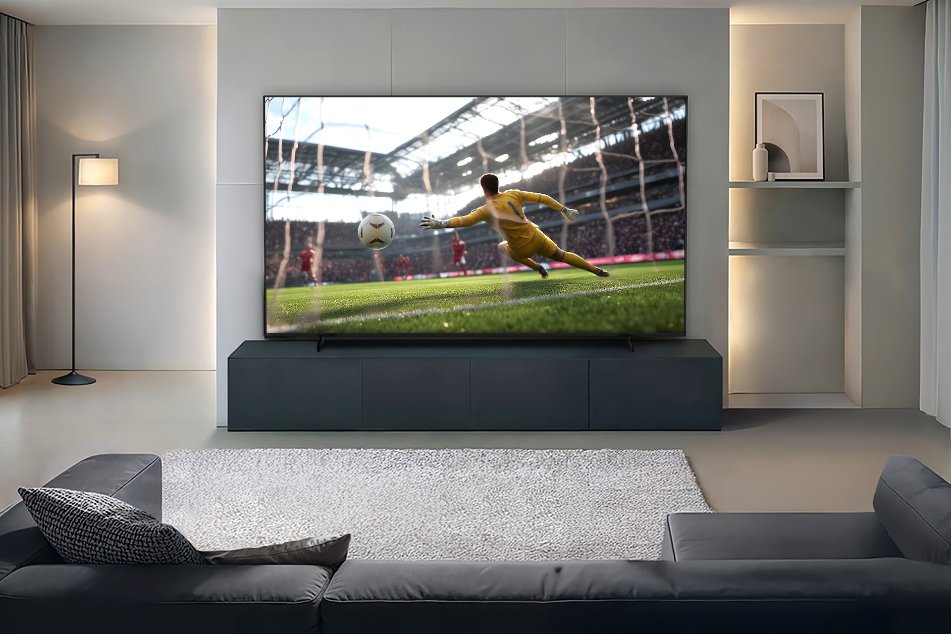거실 장식장 위에 TV가 설치되어 있습니다. TV 화면에는 축구 경기가 송출되고 있습니다. TV 앞에는 소파가 놓여있습니다. TV 오른쪽에는 장식장이 있으며, 장식장 안에는 액자와 화병이 놓여 있습니다. 왼쪽에는 스탠드 조명이 놓여 있습니다.
