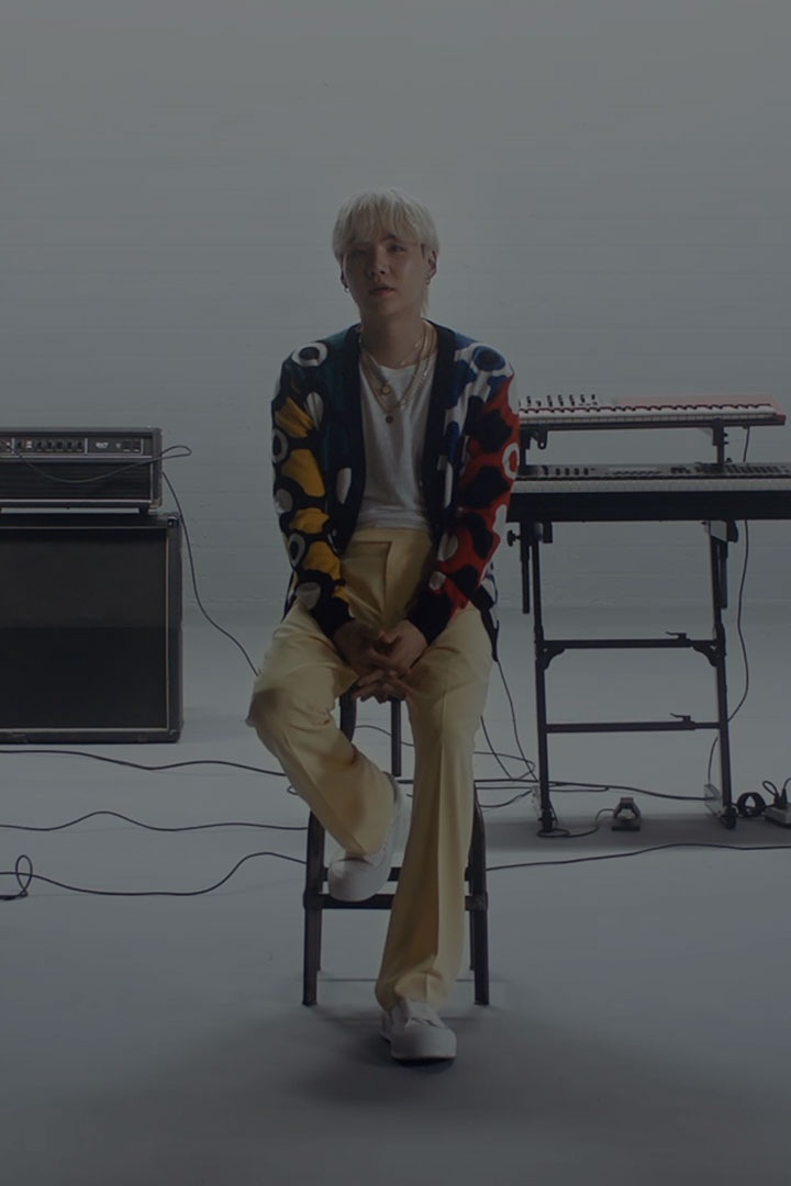 BTS 슈가가 음악 스튜디오 한가운데 의자에 앉아있습니다. 왼쪽에는 스피커와 앰프, 오른쪽에는 세 대의 전자 키보드가 있습니다.