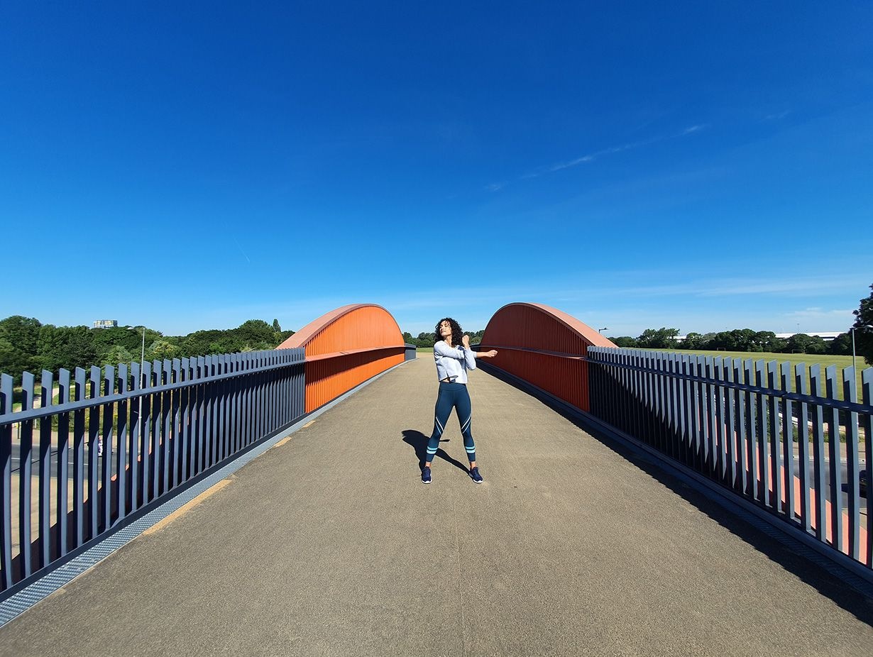 Foto tomada con cámara ultraancha de una mujer que se estira en un puente con portones grises y acentos anaranjados que contrastan con un cielo azul brillante