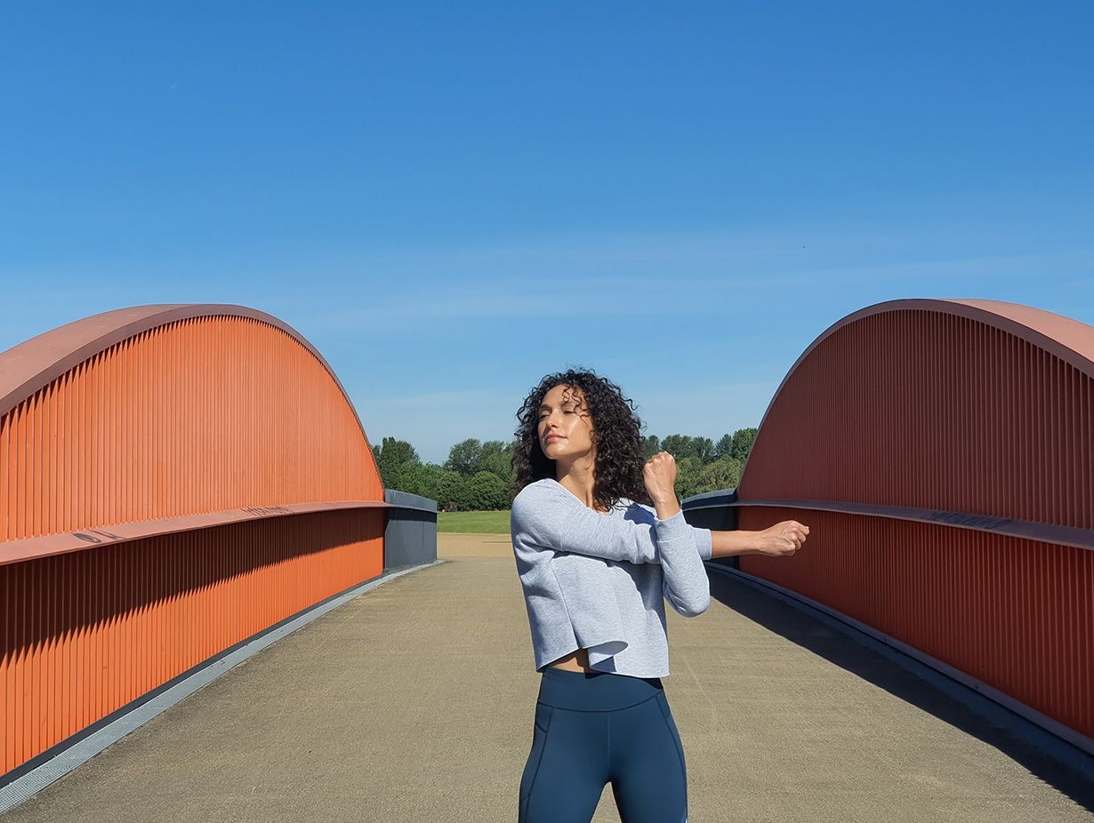 Foto tomada con cámara de telefotografía de una mujer que se estira en un puente con portones grises y acentos anaranjados que contrastan con un cielo azul brillante
