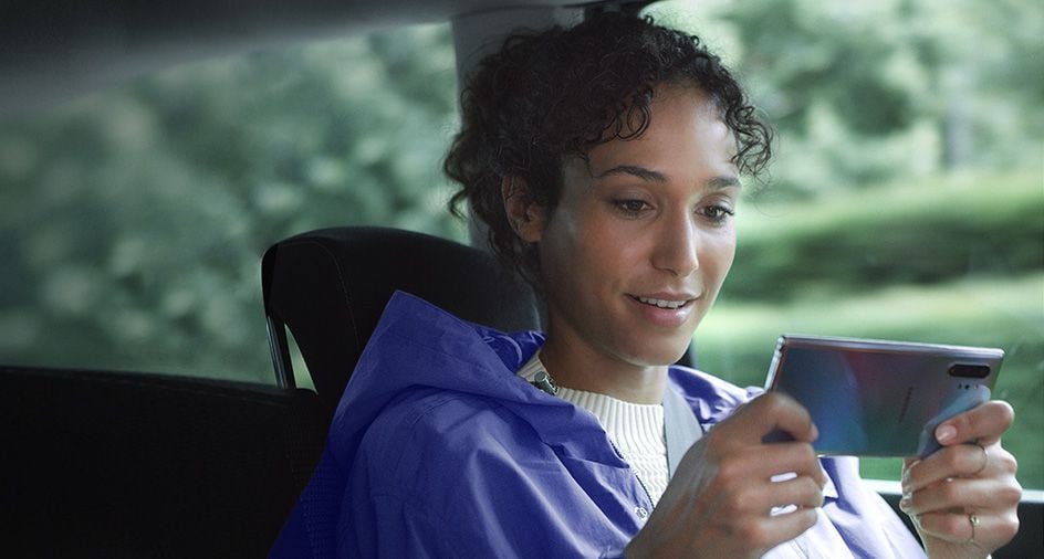 Mujer sentada en un auto sostiene un Galaxy Note10 plus en modo horizontal. Aparecen notificaciones del gestor de descargas que muestran que, gracias a las capacidades de datos rápidas, se están descargando simultáneamente tres archivos de gran tamaño