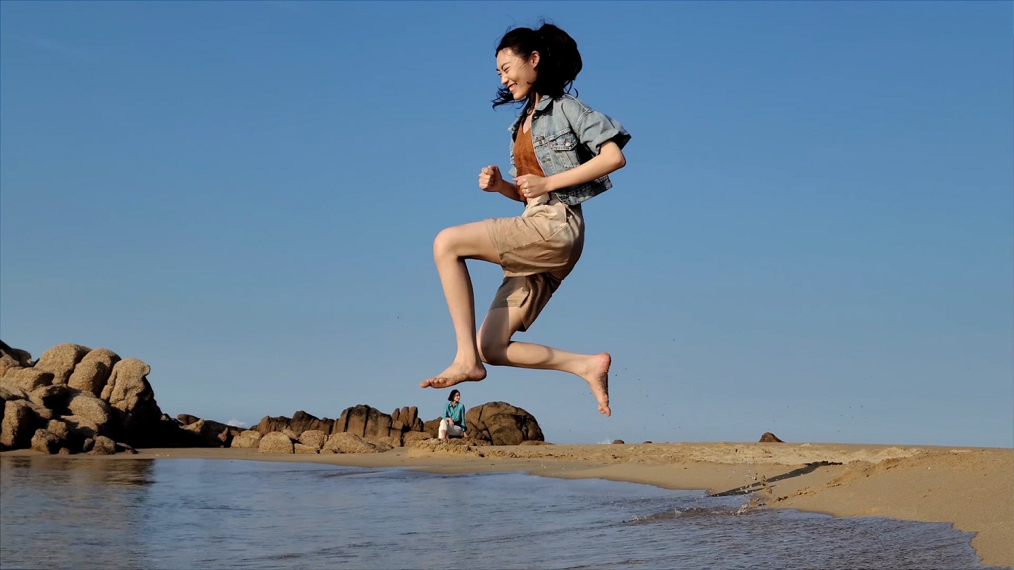 Video 8K de una mujer sentada sobre una roca en la playa y otra mujer que viene corriendo y salta al agua. La línea de tiempo se borra hasta que vemos un fotograma de la mujer saltando, y parece que tiene el pie sobre la cabeza de la otra mujer, capturando un momento divertido que de otro modo se habría pasado por alto.