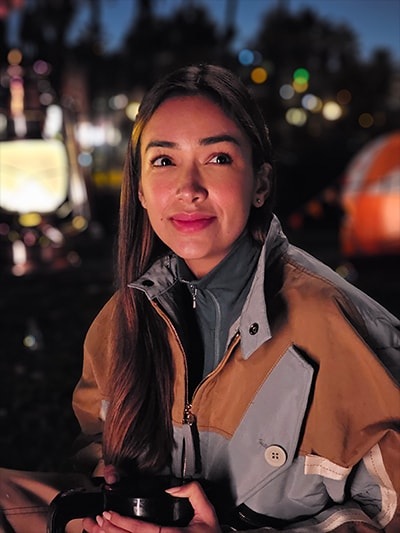 Foto retrato de cámara nocturna de una mujer sentada afuera en un campamento. Las luces de las cuerdas iluminan levemente el fondo oscuro. Luego, un retrato del mismo sujeto a una distancia adicional. Los retratos de cámara nocturna mantienen al sujeto en un enfoque claro con colores brillantes y ruido reducido en condiciones de poca luz. La primera foto se tomó con el Galaxy S23 Ultra usando ISO 3200, modo ancho y un diafragma de F 2.4. La segunda foto se tomó con el Galaxy S23 Ultra usando ISO 2000, modo ancho y un diafragma de F 1.7.