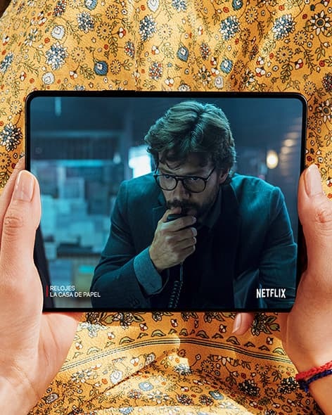 Unas manos sosteniendo el Galaxy Z Fold3 5G desplegado. La pantalla principal muestra la escena de un video de Netflix de un hombre que se lleva la mano a la cara.