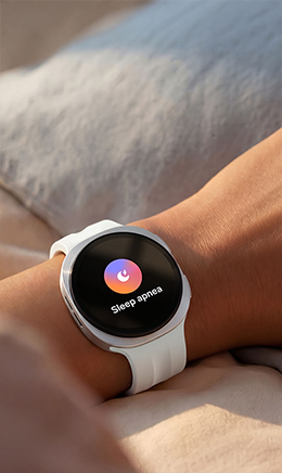 A close-up view of a hand reaching for a Galaxy smartphone on a side table facing down. On the wrist is Galaxy Watch9 in Cream with Sport Band in Cream. Sleep related feature tiles are on display on the Galaxy Watch9, indicating the person is trying to check the details of sleep tracked overnight.
