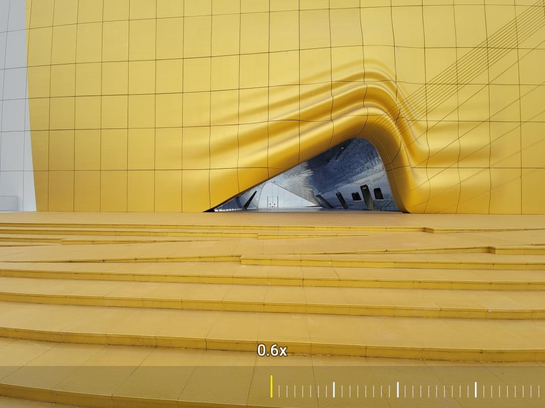 A woman seen standing on the other side of a building. As you zoom in you see she's holding an umbrella. As you zoom in further, you see her clearly. Zoom all the way in and you see a close-up of her umbrella.