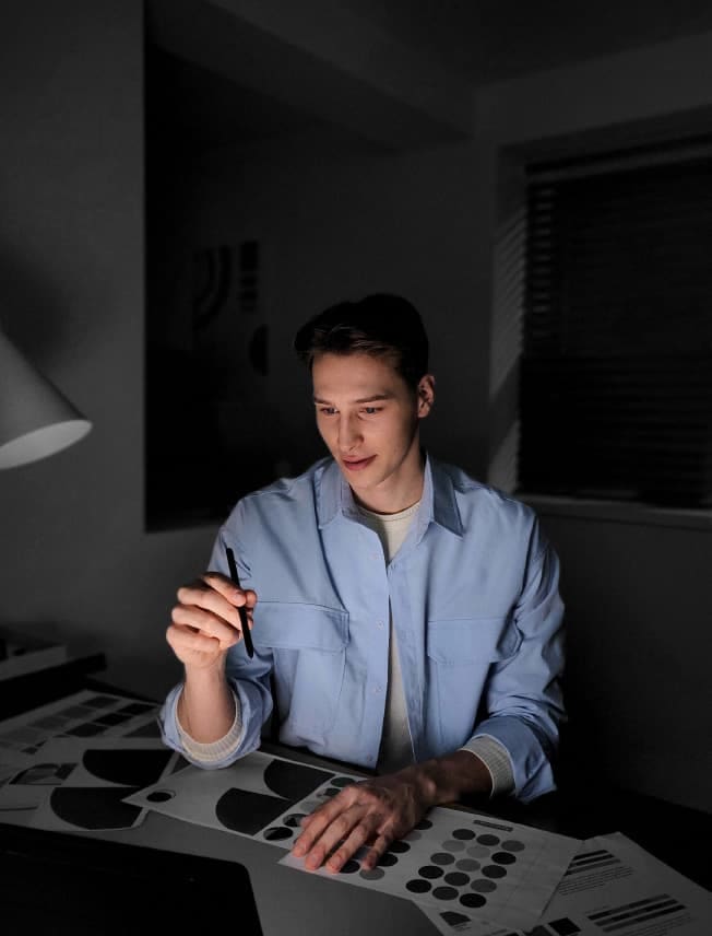 A man is sitting at a desk in a dark room with draft papers in front of him. He is holding an S Pen and looking at a device screen. His portrait is clear and bright, showcasing how Super Night Solution works to make portraits taken in darker light settings brighter and more beautiful.