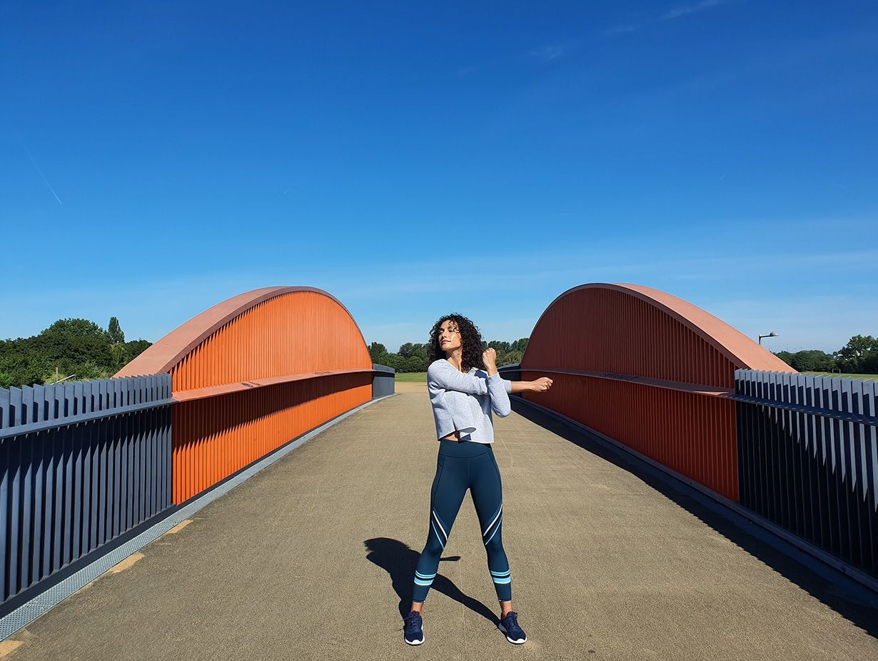Photo captured by the Wide-angle Camera of a woman stretching on a bridge with grey gating and orange accents against a bright blue sky